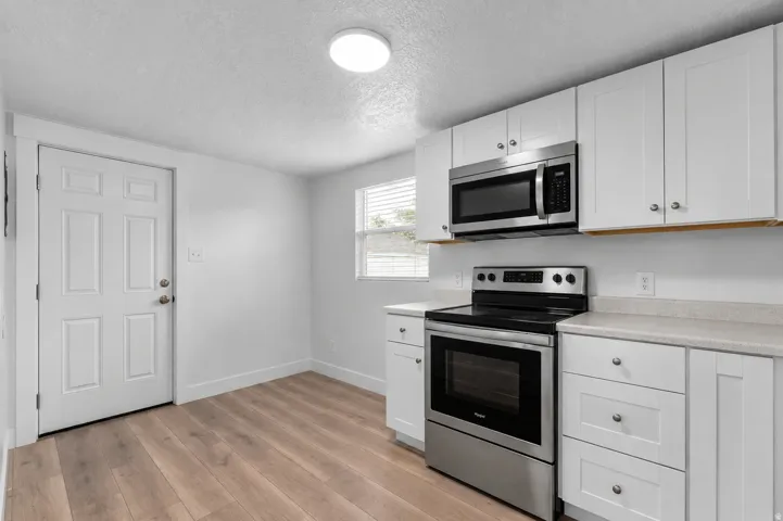 Appliances are virtually staged (actual stainless steel appliances are included). White shaker-style cabinetry, stainless steel oven/range, built-in microwave, wood-finish flooring, and a single window with blinds