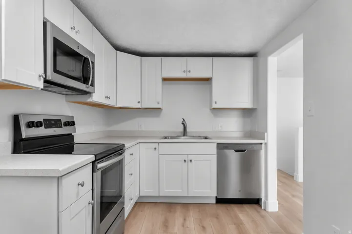Appliances are virtually staged (actual stainless steel appliances are included). White cabinetry with polished hardware, stainless steel appliances, light-toned countertops, wood-finish flooring, and a single-basin sink with a gooseneck faucet