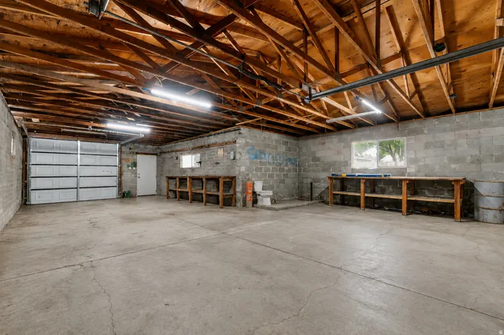 Spacious garage featuring a concrete floor, exposed wood ceiling beams, multiple fluorescent light fixtures, and a roll-up garage door