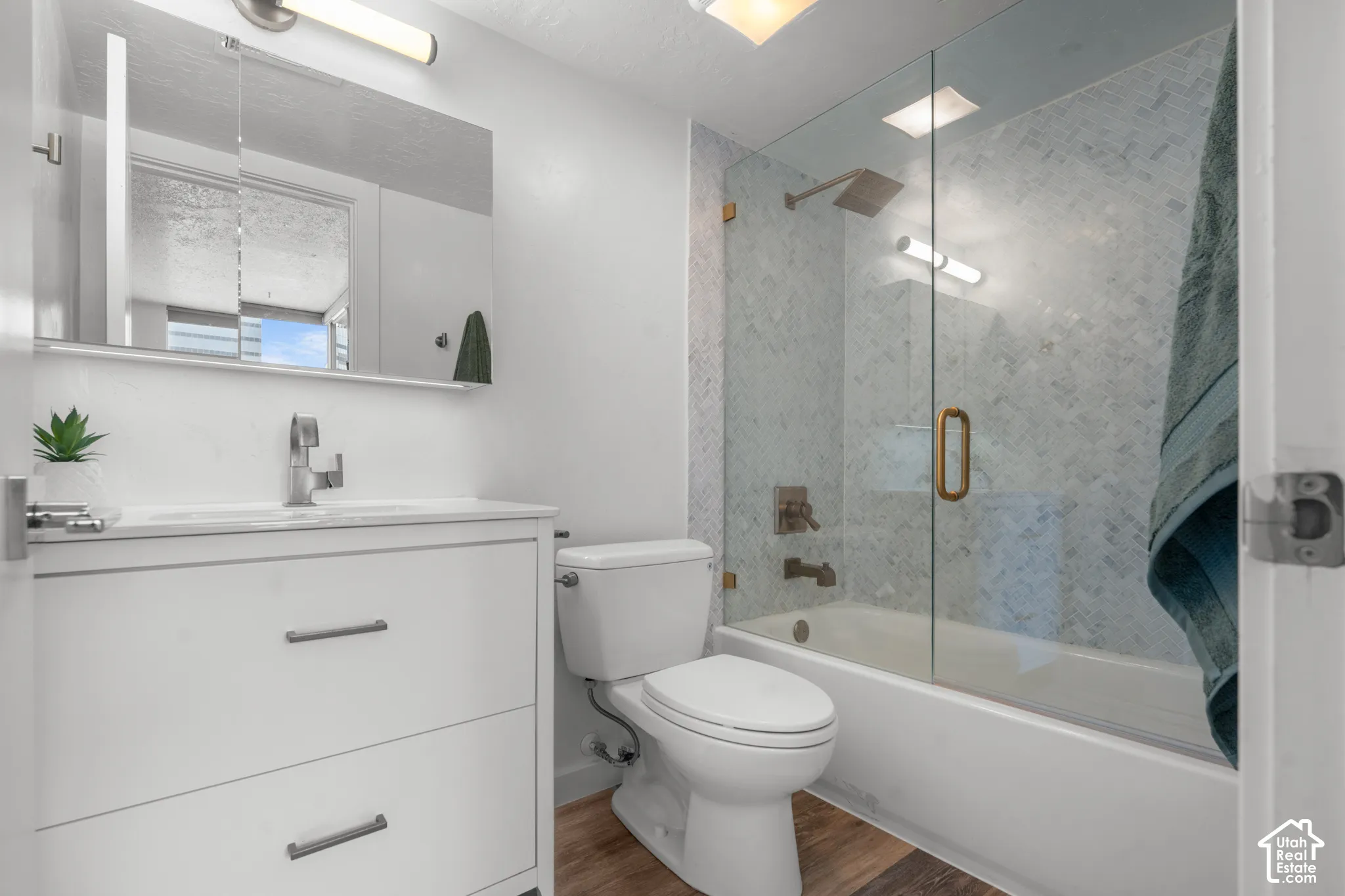 Vanity with a single basin sink, wall-mounted mirror, recessed overhead lighting, and a glass-enclosed shower-tub combination with tiled walls