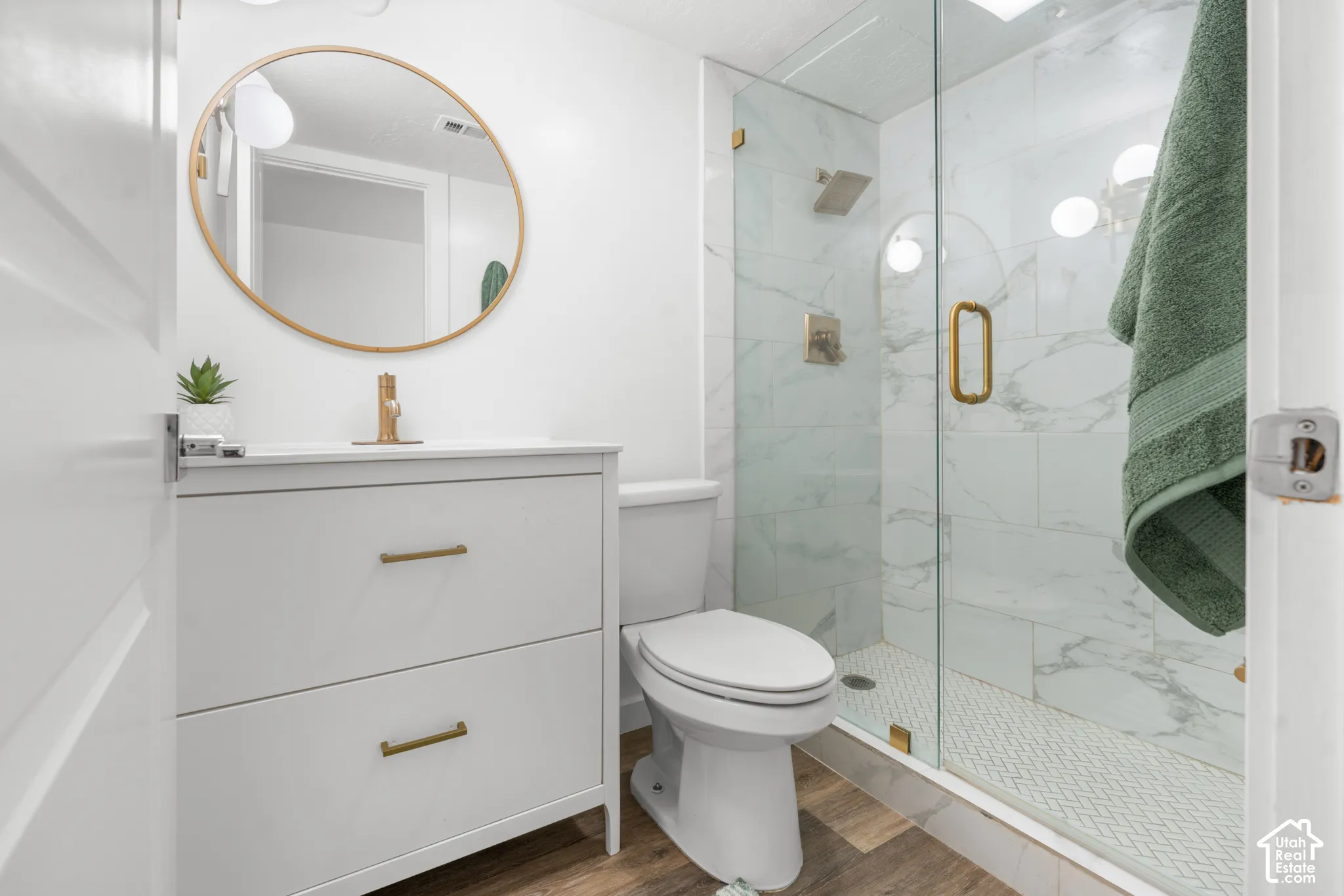White vanity with brass hardware, wood-finish flooring, glass-enclosed shower, white tiled shower surround, and gold-finish shower fixtures