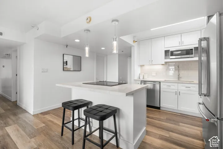 White cabinetry with stainless steel appliances, wood-finish flooring, island with integrated cooktop, and subway tile backsplash