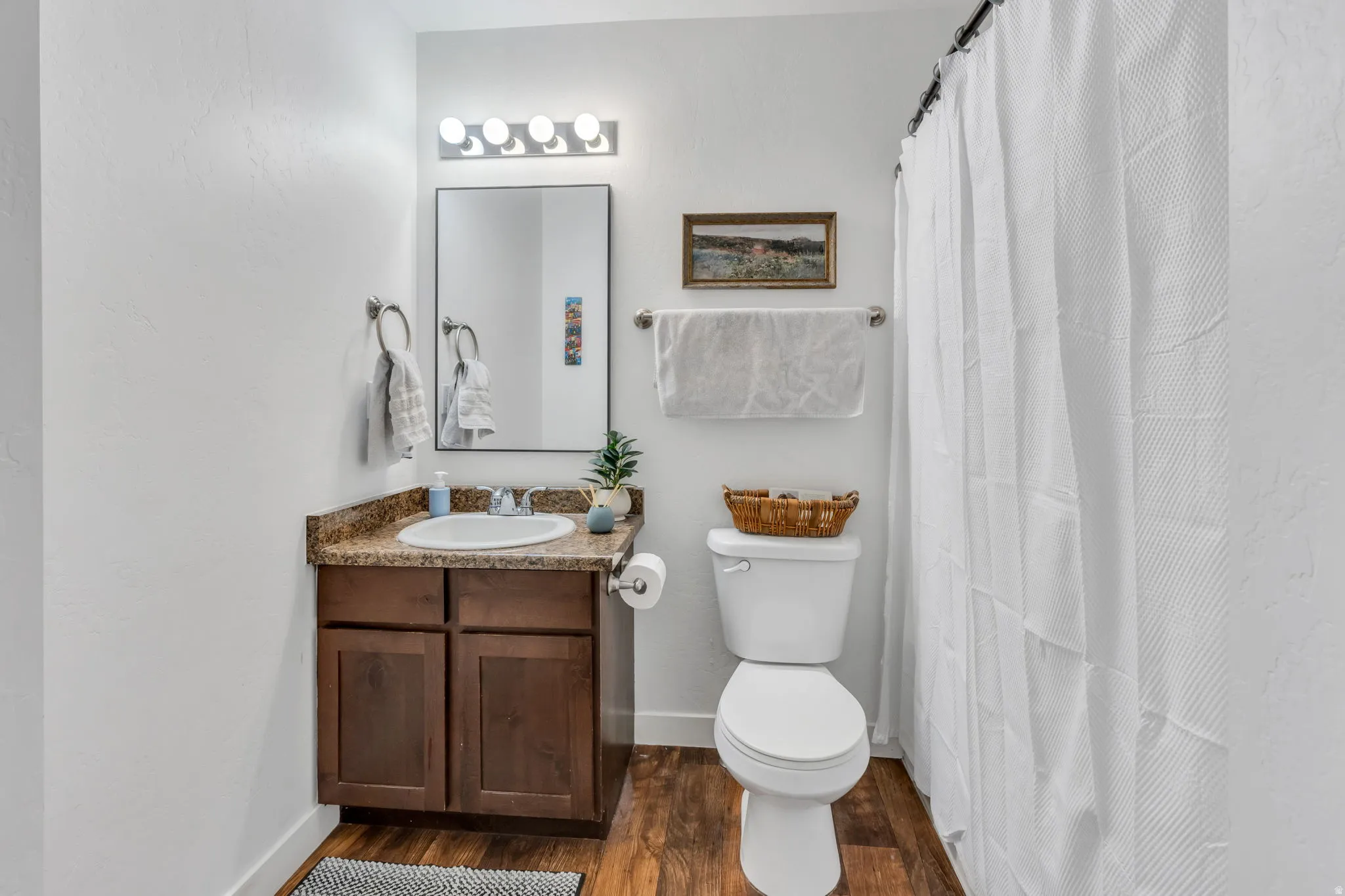 Wood-finish flooring, dark wood vanity with cabinet storage, stone-finish countertop, single basin sink, and a wall-mounted mirror