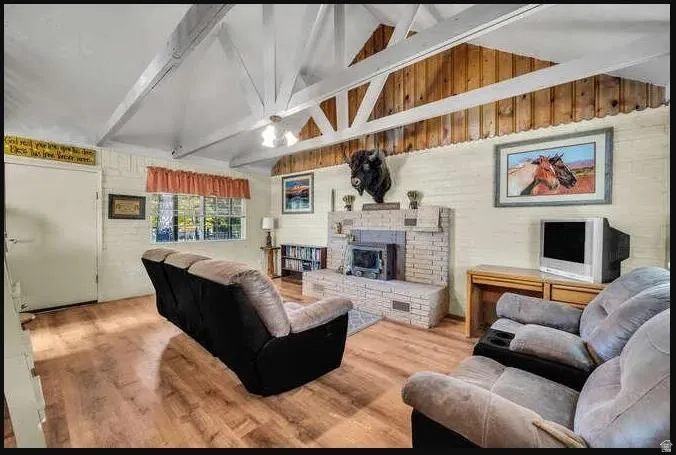 Vaulted ceiling with exposed white beams and wood plank accents, wood-finish flooring, brick-faced fireplace, built-in shelving, and multiple windows