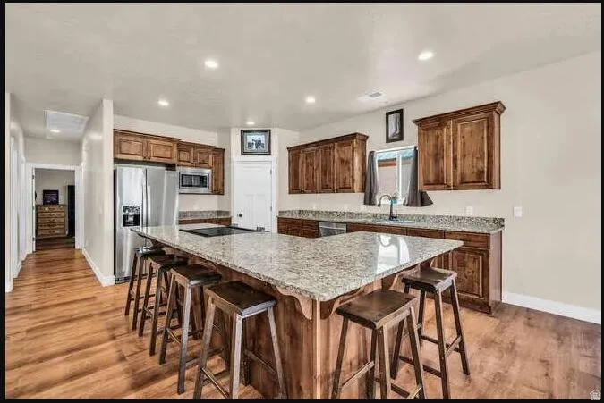 Spacious kitchen featuring a large L-shaped island with integrated cooktop, wood-finish flooring, recessed lighting, wood cabinetry, and stainless-steel appliances