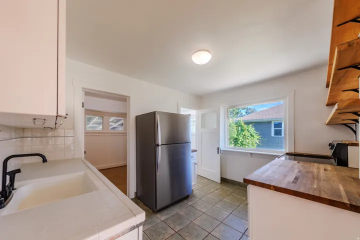 White cabinetry, tile backsplash, wood-finish countertops, stainless steel refrigerator, and tiled flooring