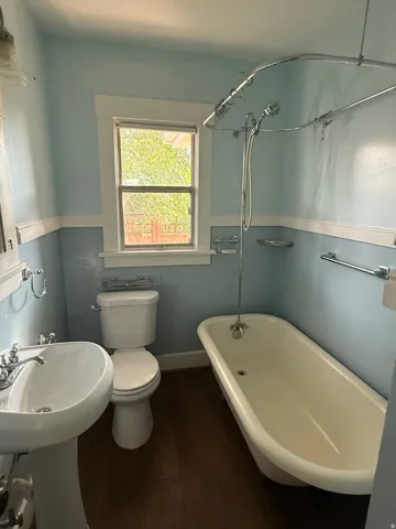 Pedestal sink with chrome fixtures, wood-finish flooring, white clawfoot tub with shower attachment, and a window with natural light