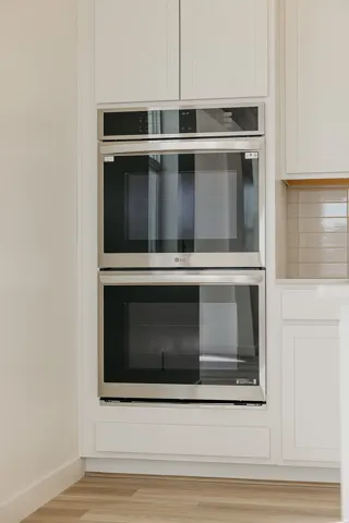 Stainless steel double wall oven with integrated microwave, surrounded by white shaker-style cabinetry and wood-finish flooring