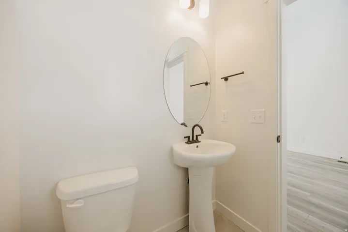 Pedestal sink with dark bronze faucet, oval mirror, wall-mounted light fixture, and tile flooring