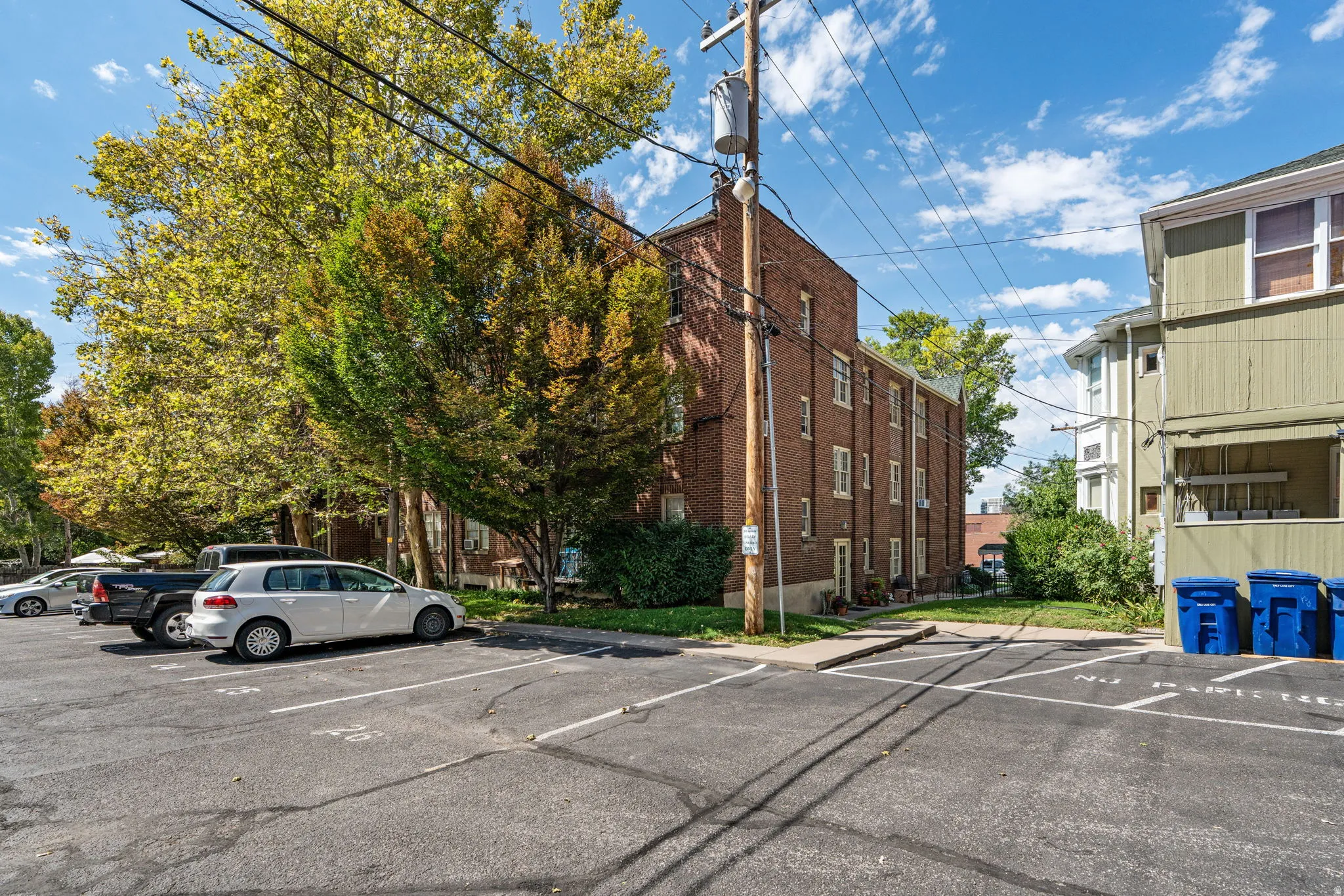Brick-clad building with multiple windows, surrounded by mature trees, and an asphalt parking area