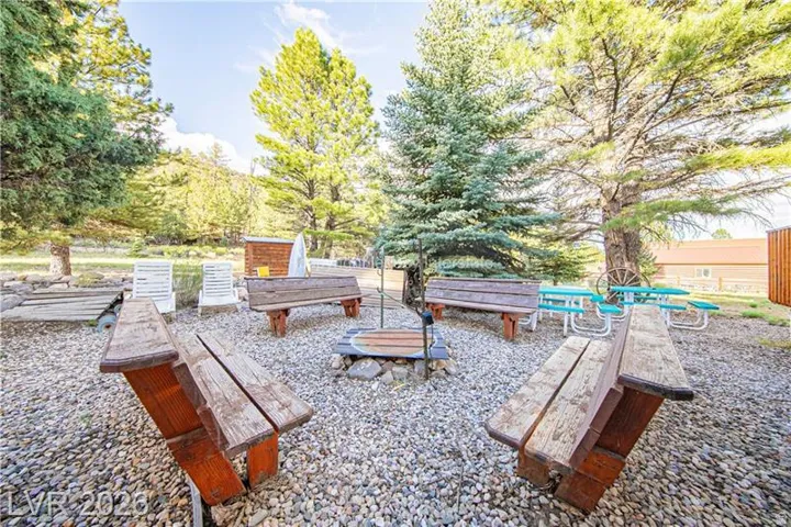 Gravel-covered outdoor area with a central fire pit, surrounded by multiple wooden benches and mature trees