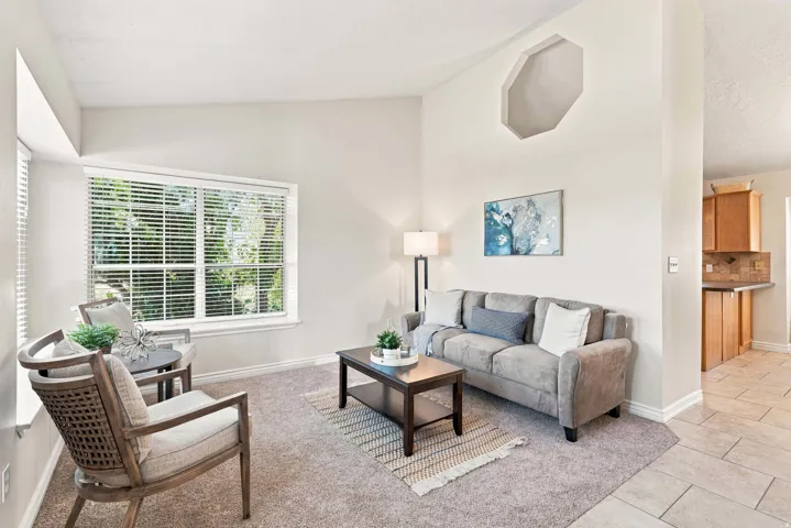 Vaulted ceilings with architectural detail, large window with blinds, light-toned carpet, tiled flooring, and a partial view of a kitchen with wood cabinetry