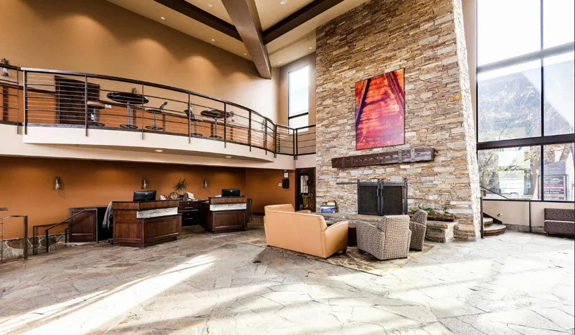 Spacious common area featuring a stone accent wall fireplace, high ceilings with exposed beams, expansive window walls, an upper-level balcony with metal railing, and stone tile flooring