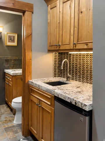 Wood cabinetry with integrated under-cabinet lighting, granite-style countertop, stainless steel bar sink, and mosaic tile backsplash