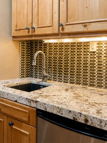 Wood cabinetry with dark hardware, stone countertop, stainless steel sink, pull-down faucet, and patterned mosaic tile backsplash