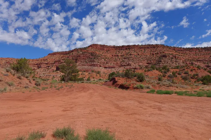 Expansive red earth terrain with a visible track, featuring scattered desert vegetation and towering rock formations