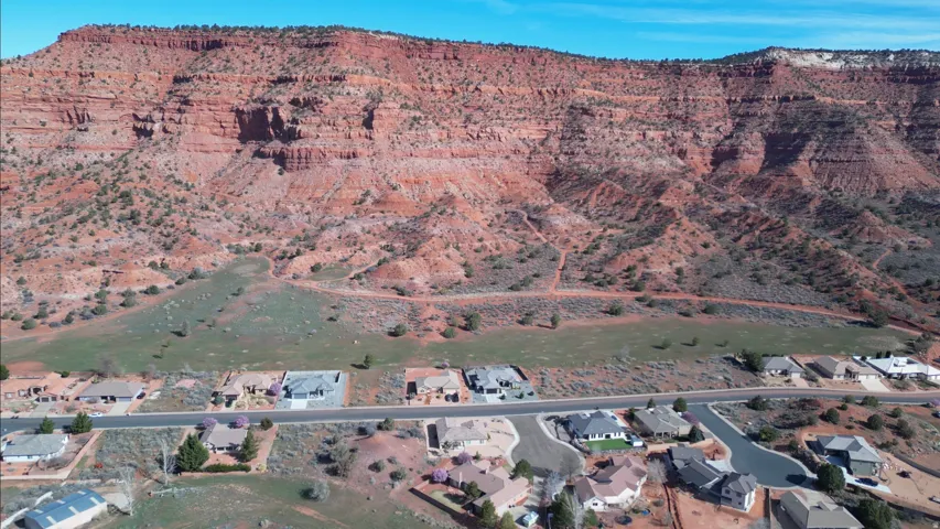 Red rock mountain backdrop, residential street with varying home styles, green natural terrain, and clear blue sky
