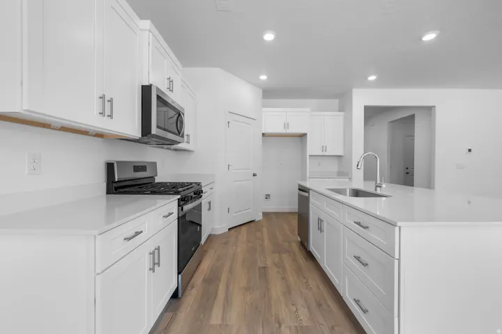 Modern galley kitchen featuring white shaker cabinetry, stainless steel appliances including a gas range and microwave, quartz countertops, brushed nickel hardware, recessed lighting, and warm wood-finish flooring throughout