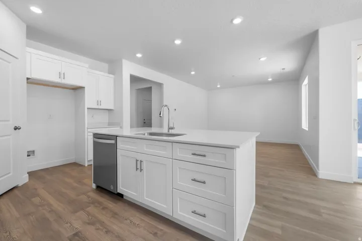 Kitchen featuring an island with a built-in sink and dishwasher, white shaker-style cabinetry, light-toned countertops, and wood-finish flooring