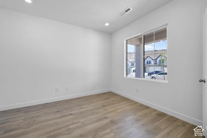 Bright, light-filled bedroom showcasing warm wood-finish flooring, recessed ceiling lighting, and a large picture window with natural daylight