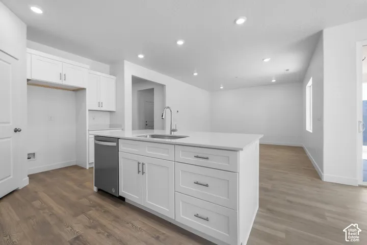 Kitchen featuring an island with a built-in sink and dishwasher, white shaker-style cabinetry, light-toned countertops, and wood-finish flooring