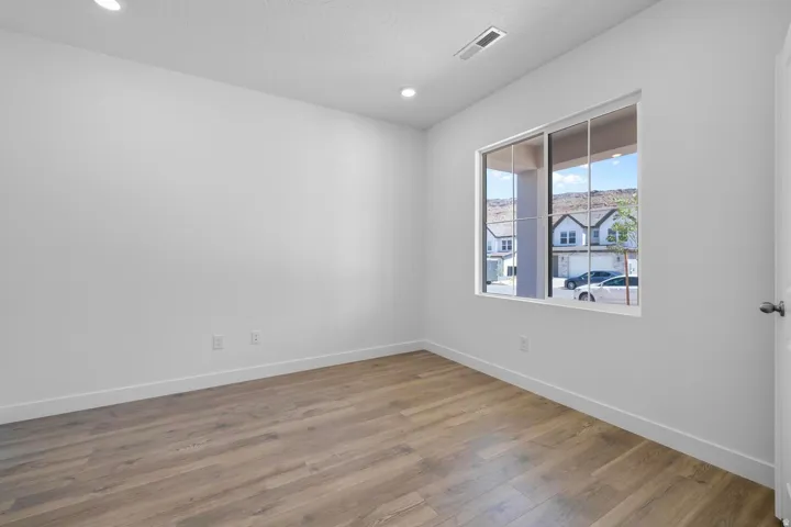 Bright, light-filled bedroom showcasing warm wood-finish flooring, recessed ceiling lighting, and a large picture window with natural daylight