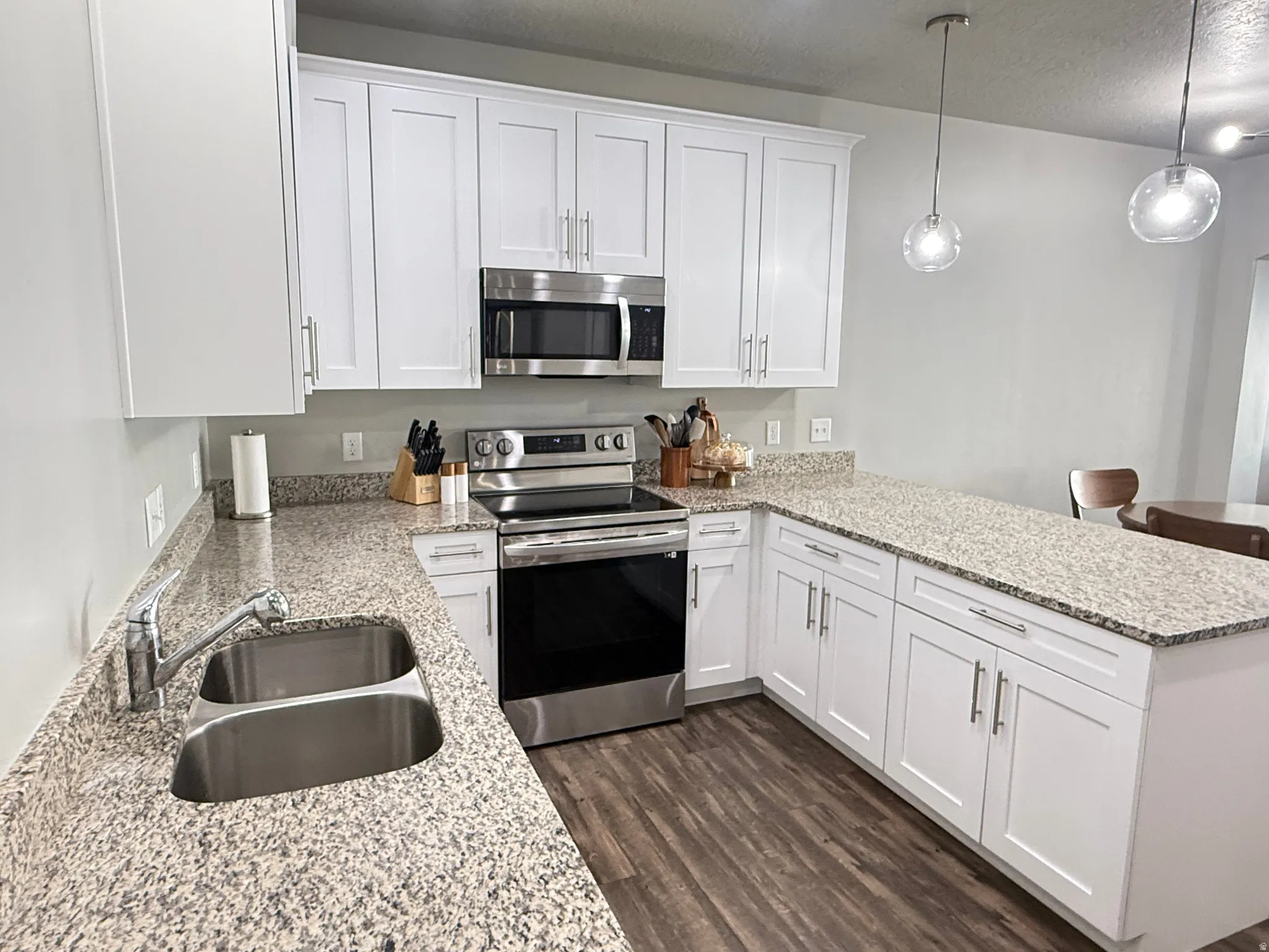 Contemporary kitchen featuring white cabinetry, granite countertops, stainless steel appliances, wood-finish flooring, and pendant lighting