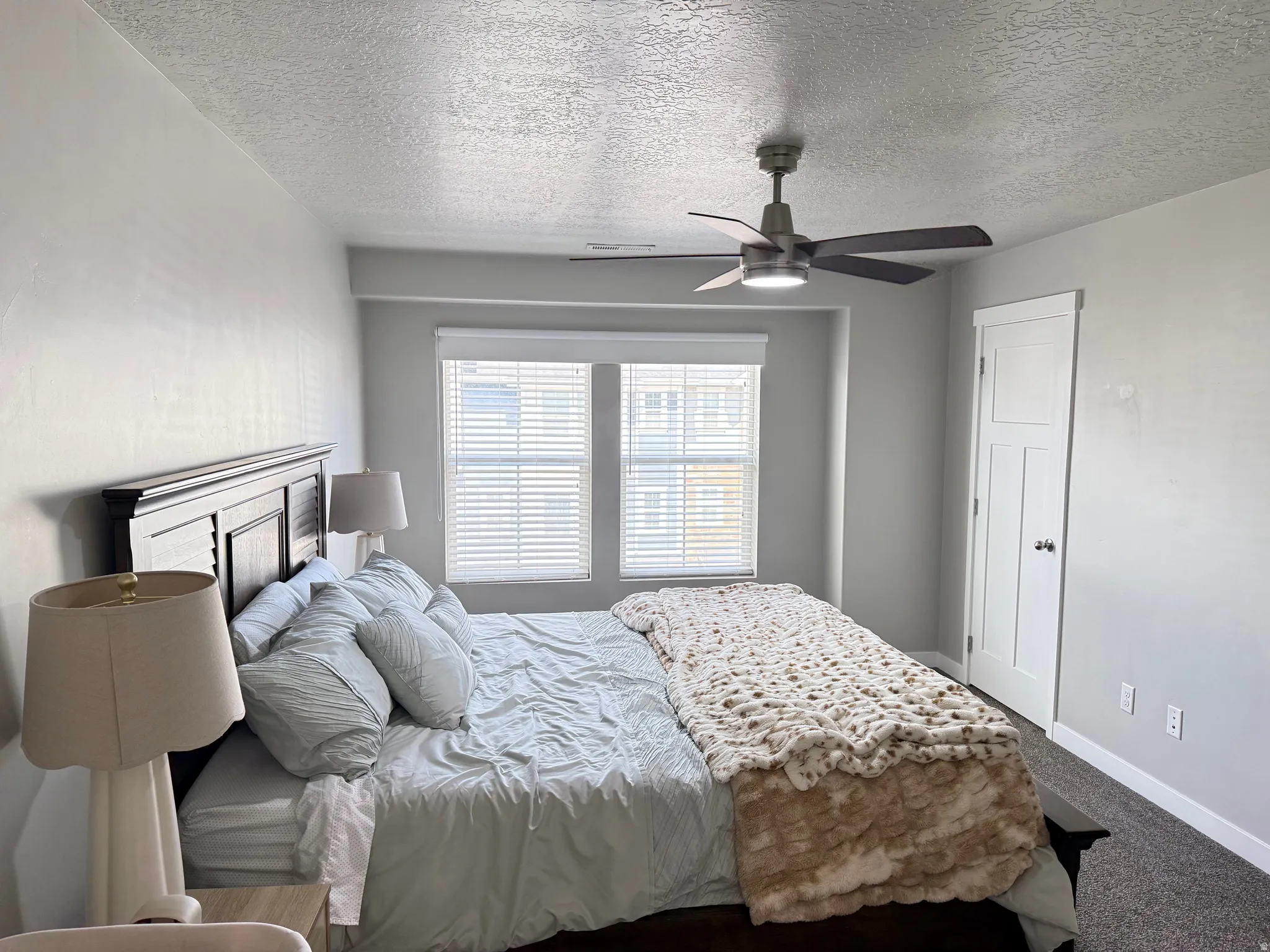 Recessed window with blinds, dark wood-finish headboard, overhead ceiling fan with lighting, white interior door, and dark carpet flooring