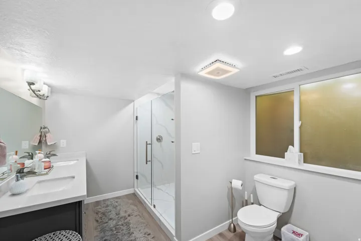 Dual sink vanity with white countertop, walk-in shower with glass enclosure, wood-finish flooring, and recessed lighting