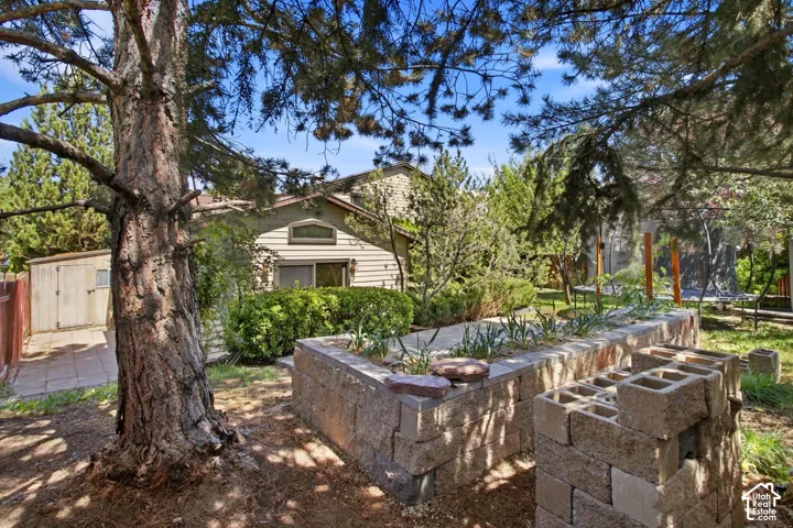 Mature trees, raised block planters, paved pathway, green shrubbery, and a storage shed