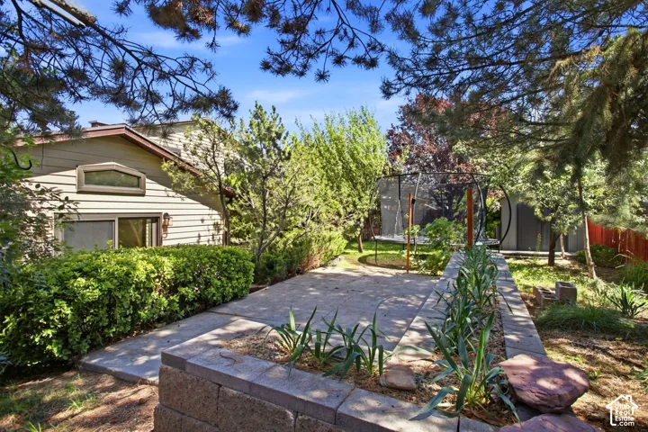 Exterior siding with a geometric window, mature perimeter landscaping, concrete patio, and garden beds with stone block retaining walls