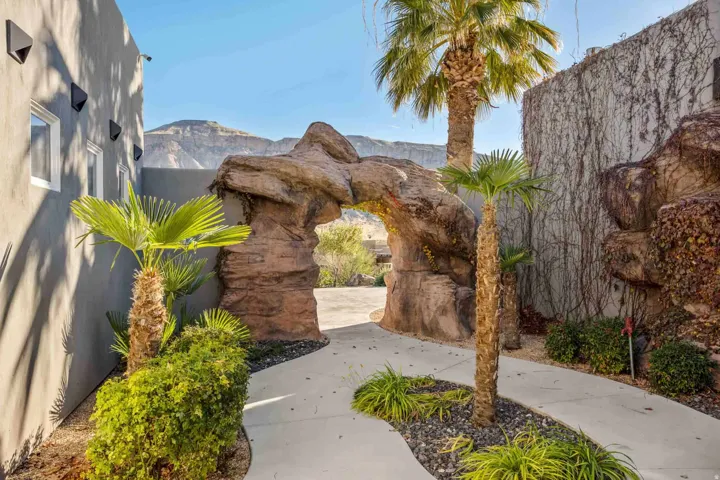 Curved concrete pathway leading to a rock archway, flanked by mature palm trees and low-lying shrubs