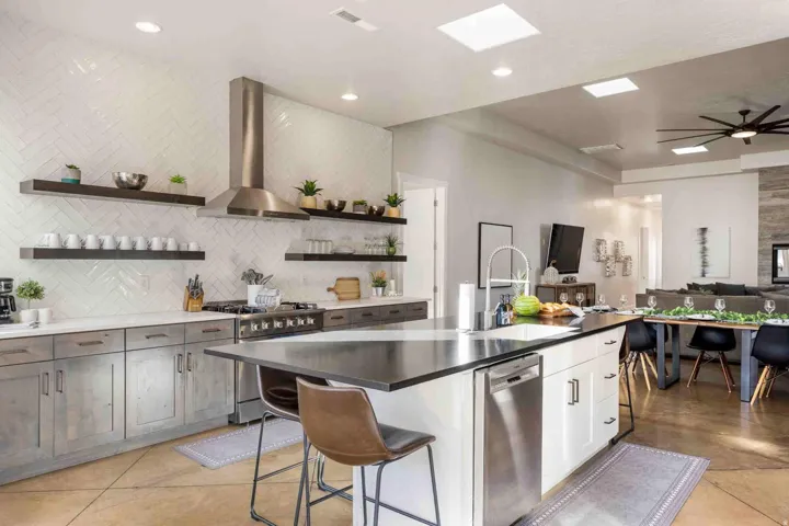 Open-concept kitchen featuring an island with built-in sink and seating, stainless steel appliances, herringbone tile backsplash, and floating shelves