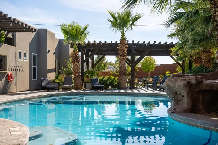 Swimming pool with integrated rock feature, surrounded by paver decking, mature palm trees, and a dark wood pergola