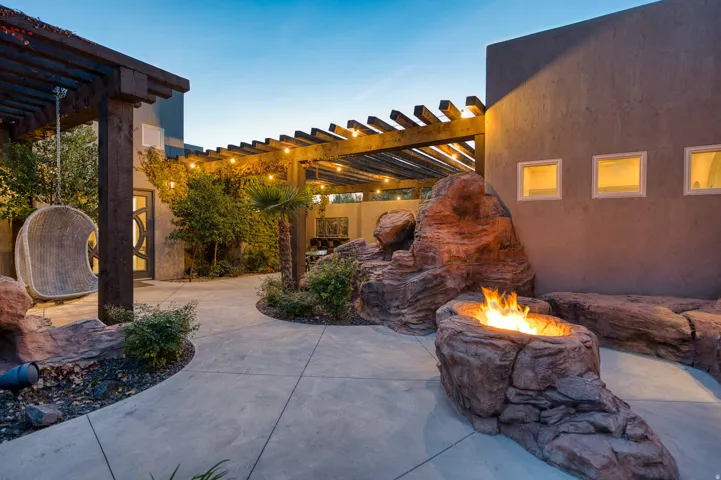 Paved courtyard featuring a rock-built fire pit, wood pergola, string lighting, and stucco exterior walls