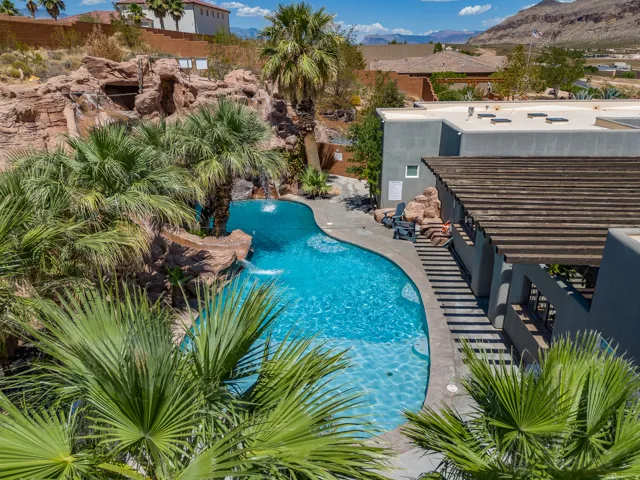 Curved swimming pool featuring rock formations, water features, mature palm trees, and a shaded pergola