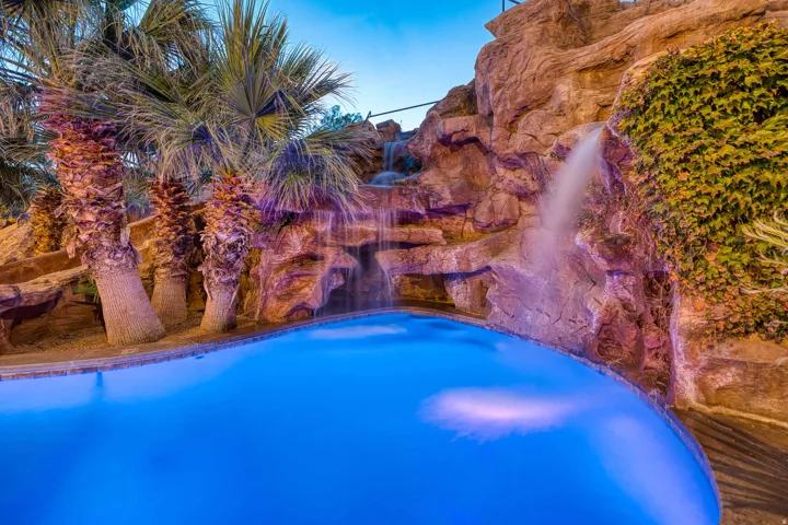 Curved pool with a rock waterfall feature, integrated lighting, and multiple palm trees