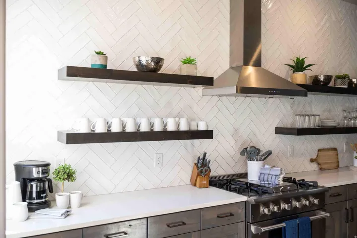 Herringbone tile backsplash, stainless steel range hood, dark wood-finish open shelving, stainless steel range, and light-toned countertops