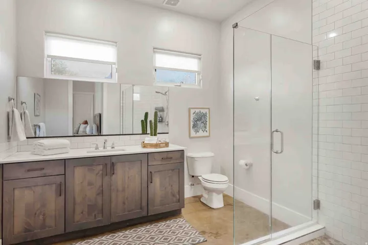 Expansive vanity with wood-finish cabinetry and white countertop, integrated sink with chrome faucet, large frameless mirror, and glass-enclosed shower with white subway tile surround