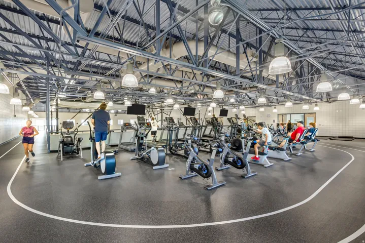 Fitness center with exposed structural ceiling, ample overhead lighting, rubber flooring with track markings, and multiple exercise machines