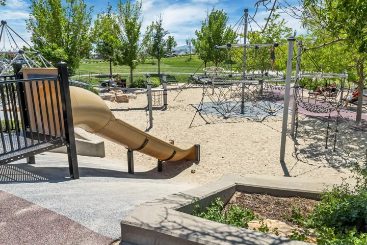 Community playground featuring a tubular slide structure, climbing apparatus, and sand surface, surrounded by mature landscaping and open green space with shade trees throughout the park grounds