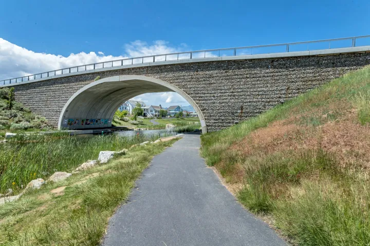 Paved pathway alongside a waterway, bordered by grassy banks and a stone-clad bridge with a concrete arch