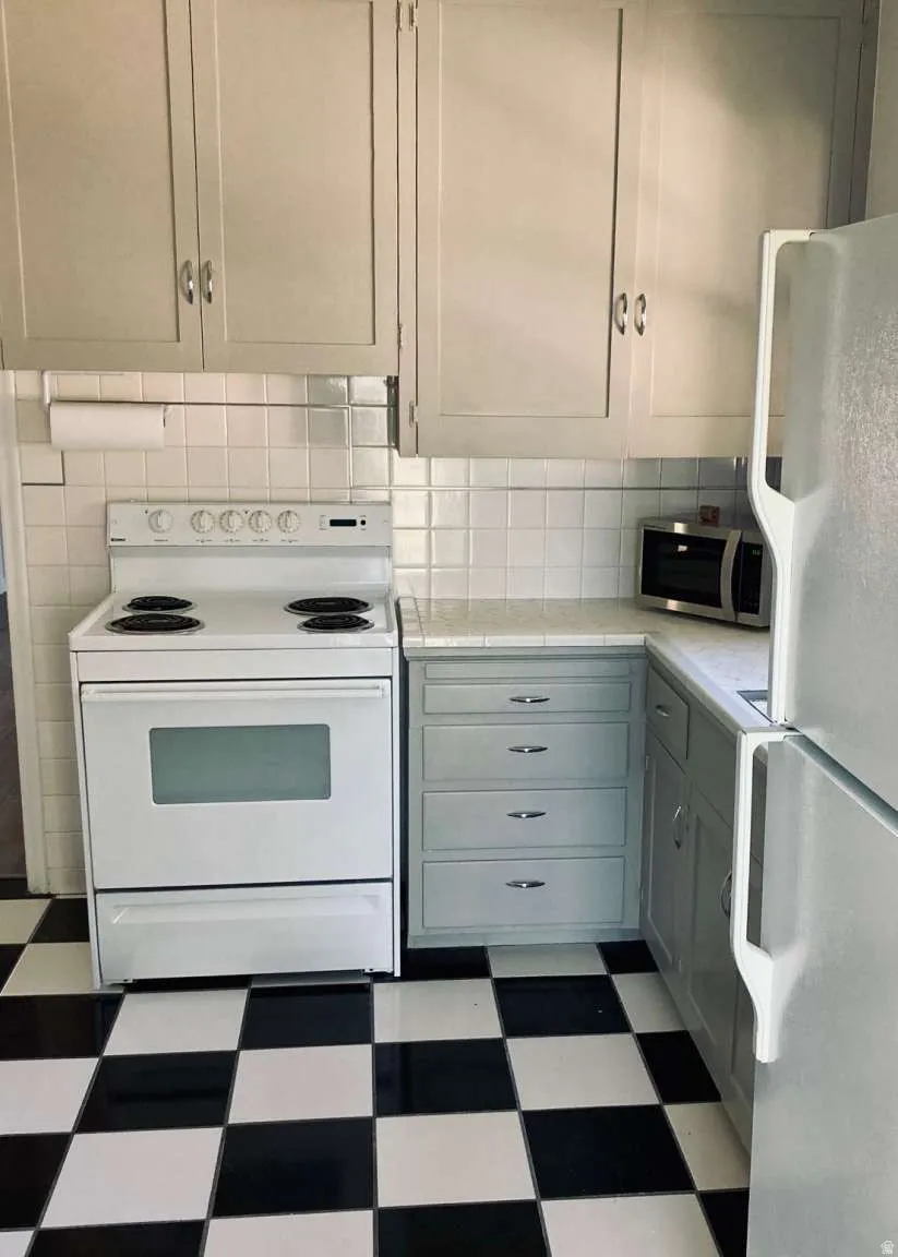 White tile backsplash, light-toned upper cabinetry, white electric range, light-toned lower cabinetry, and checkerboard floor tiling