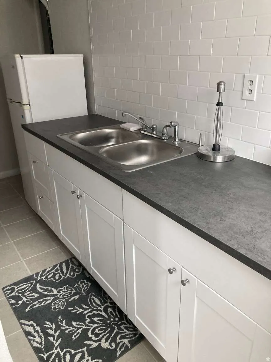 White subway tile backsplash, dual basin stainless steel sink, dark-toned countertop, white cabinetry with silver-toned hardware, and light-toned tile flooring