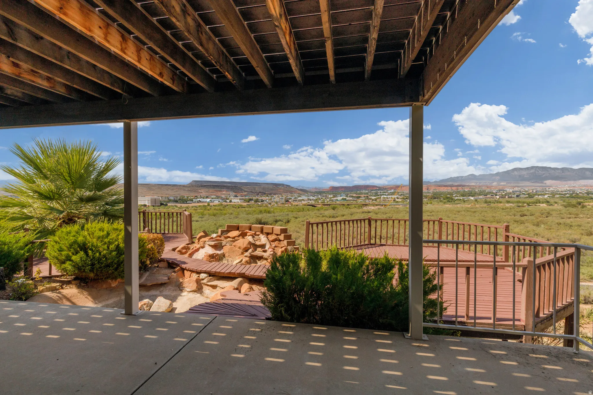 Concrete patio with overhead wood beam structure, leading to a multi-level wood deck, and a rock-lined outdoor feature with integrated wood seating