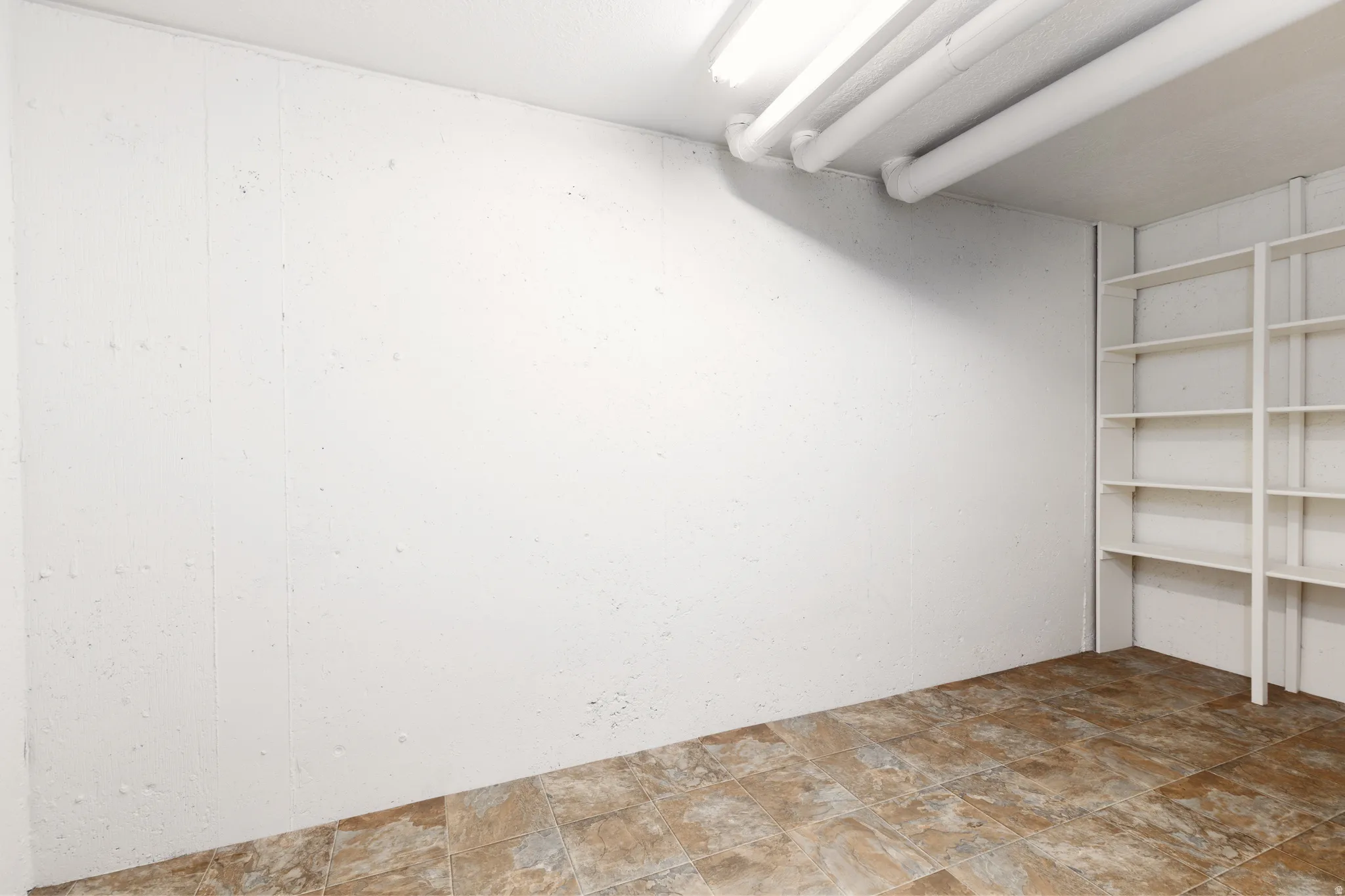 Second storage are with wall-mounted built-in shelving, tiled flooring, exposed ceiling pipes, and fluorescent lighting