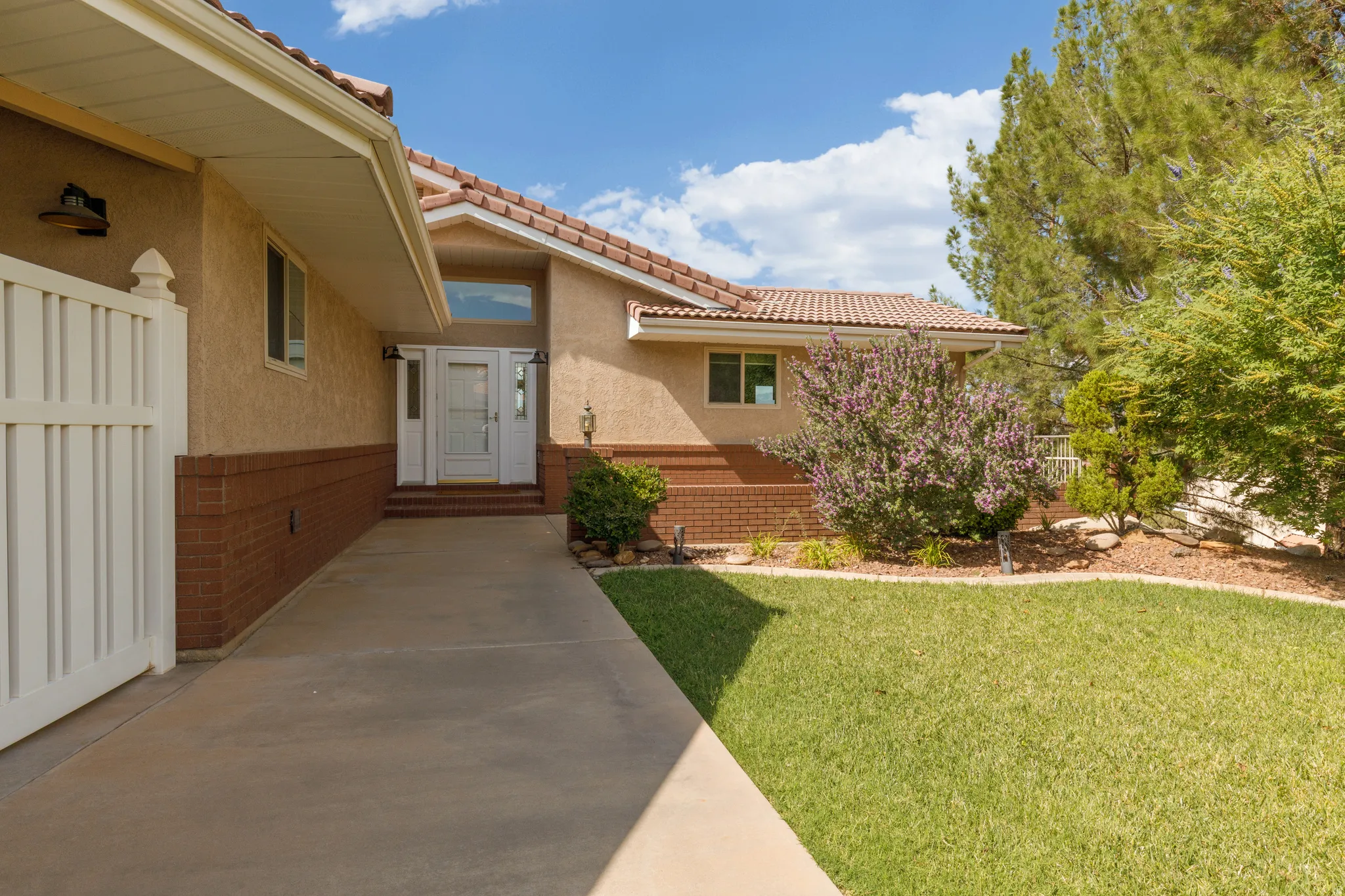 Stucco exterior with brick detailing, tile roofing, concrete walkway, white entry door with side lights, and mature landscaping