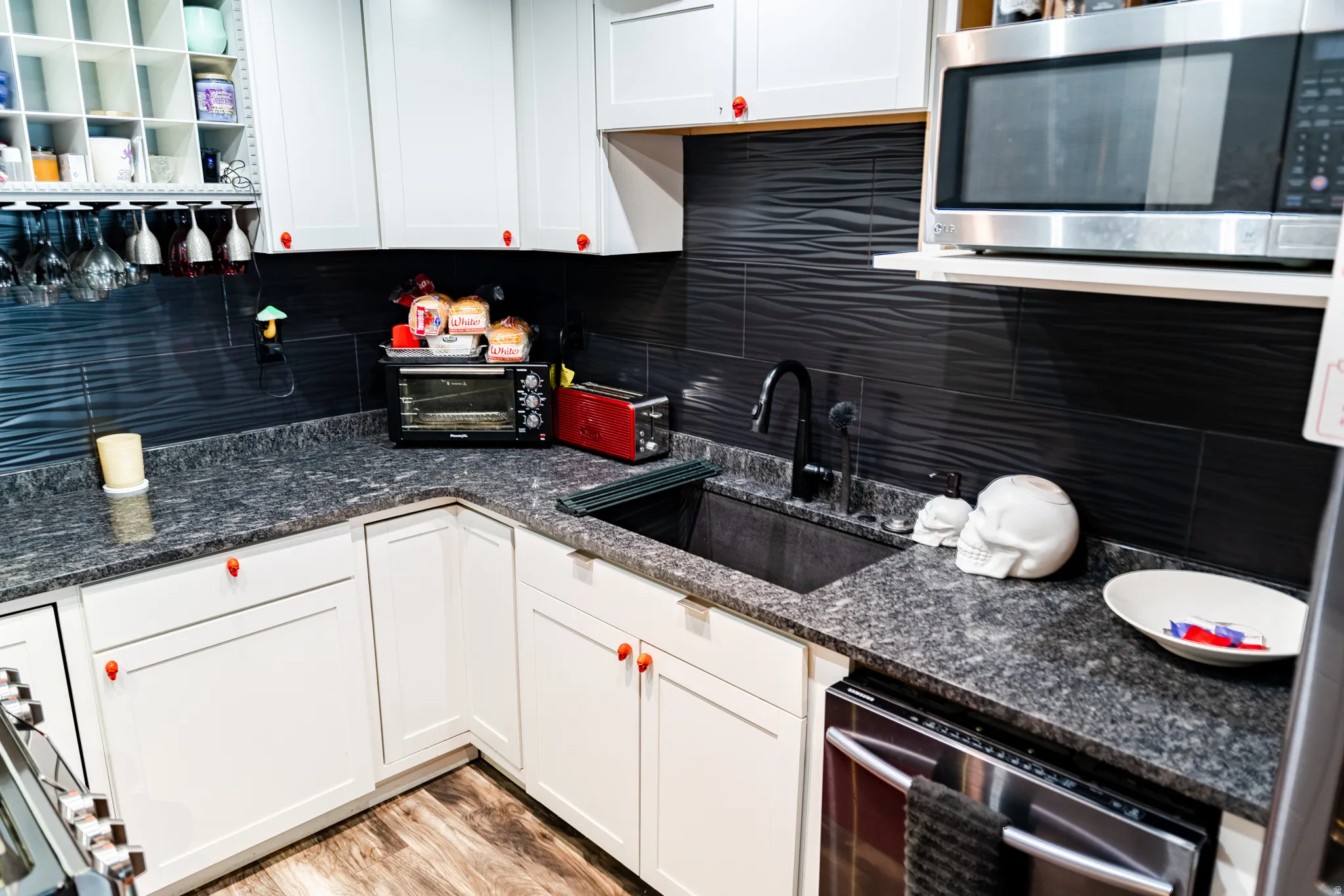 White cabinetry with red hardware, stone-finish countertops, textured dark backsplash, undermount sink with a matte black faucet, and wood-finish flooring