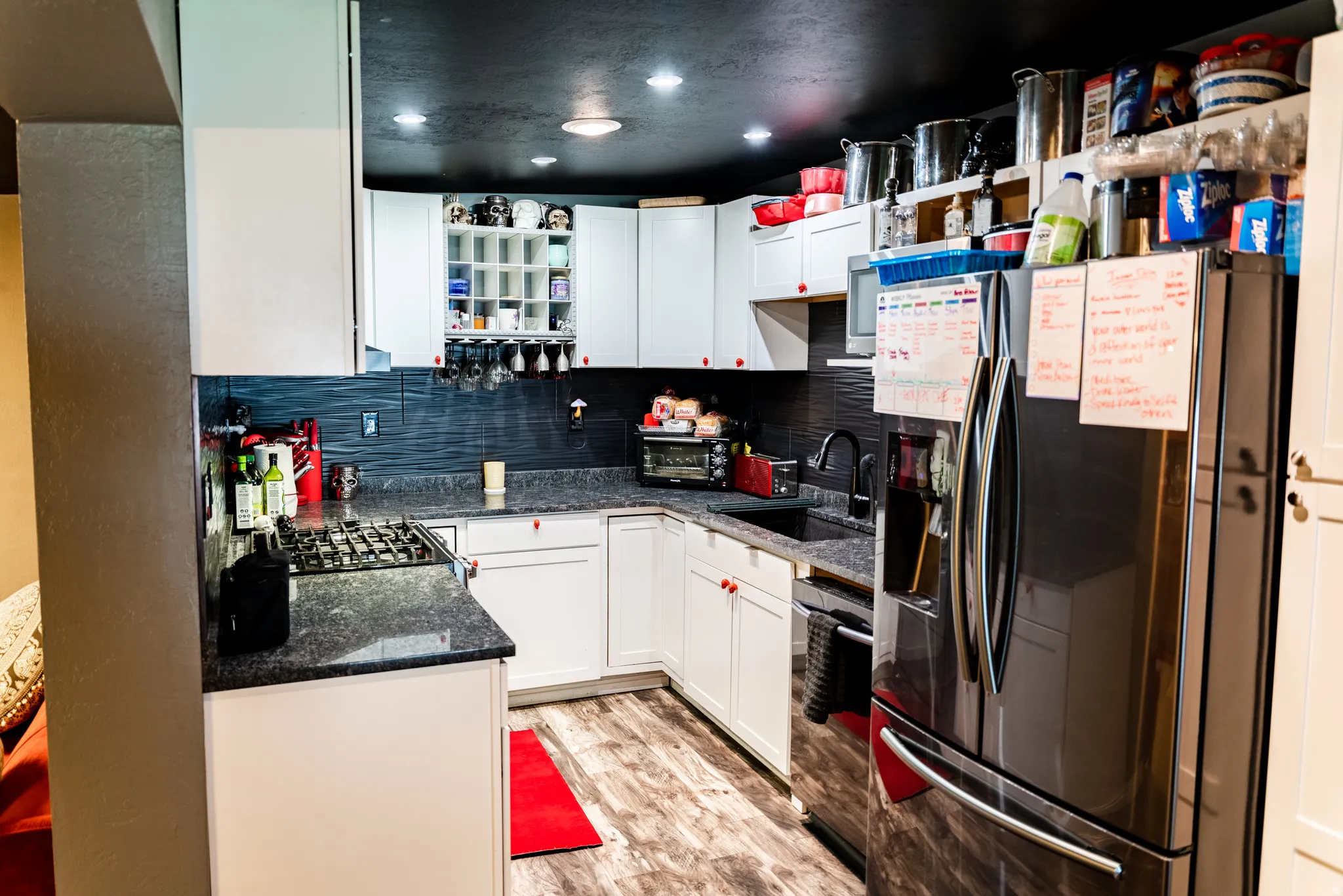 White cabinetry with red knob accents, dark textured backsplash, built-in wine storage, black stone-finish countertops, and wood-finish flooring