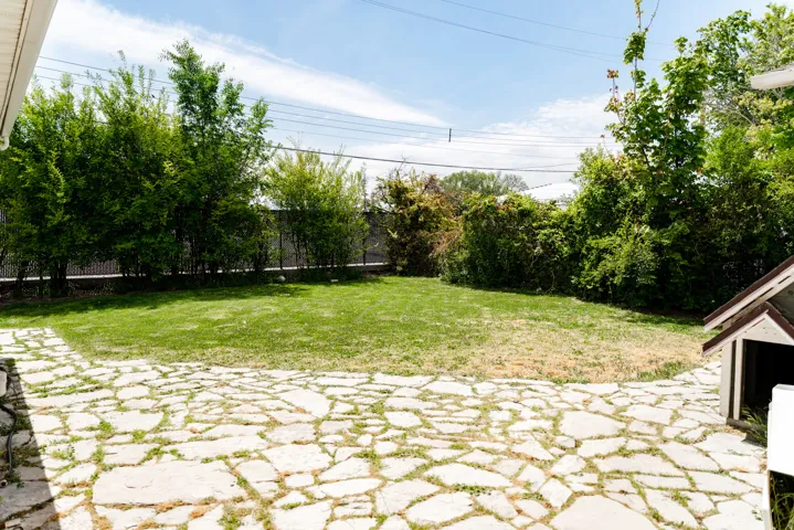 Expansive backyard featuring a natural stone patio, manicured lawn, perimeter fencing, and mature trees